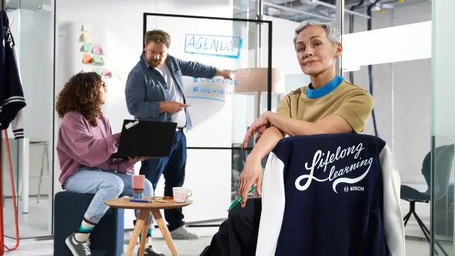 Uma senhora sentada a olhar para a câmara com o casaco "Lifelong learning", outra senhora sentada com o computador e um senhor a falar com ela enquanto aponta para um quadro que diz "Agenda; deep dive; review".