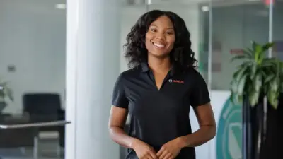 Uma jovem mulher sorridente, com uma camisa polo preta com um logotipo, está num espaço de escritório moderno e luminoso.