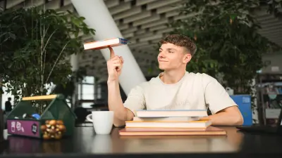 Um jovem sorri, equilibrando um livro no dedo numa mesa com mais livros e uma chávena de café num espaço moderno e arejado.