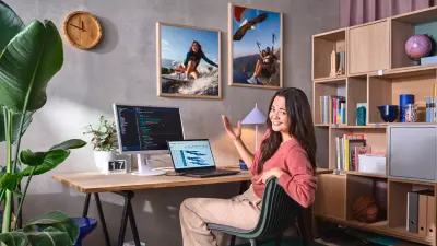 Uma jovem sorri num escritório doméstico elegante, com um portátil e um monitor a exibir código, além de decorações pessoais.