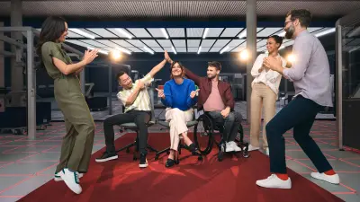 Um grupo diversificado de seis jovens celebra num tapete vermelho num espaço de escritório moderno de estilo industrial. Um homem está numa cadeira de rodas.